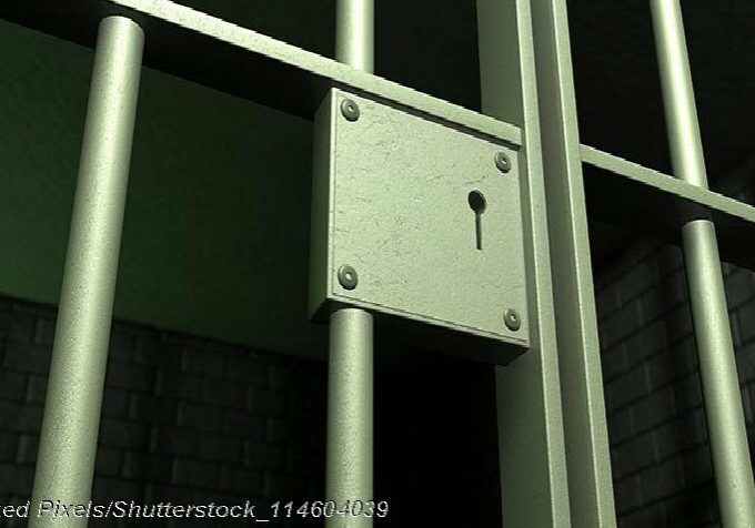 A closeup of the lock of a brick jail cell with iron bars and a door with a locking mechanism - Photo: © Copyright Linked Pixels/Shutterstock